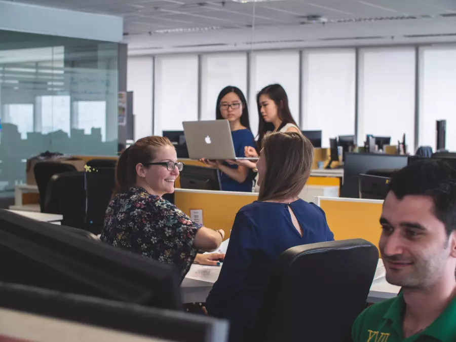 Mehrere Personen sitzen in einem modernen Büro, wobei zwei Frauen an Computern arbeiten und eine Frau im Hintergrund einen Laptop hält.